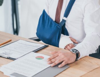 cropped view of worker with broken arm in bandage sitting at table in office, compensation concept